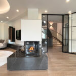 Modernes Wohnzimmer mit Kamin, Lounge-Sofas, Glasdrehtür und beleuchteter Wendeltreppe im Hintergrund.